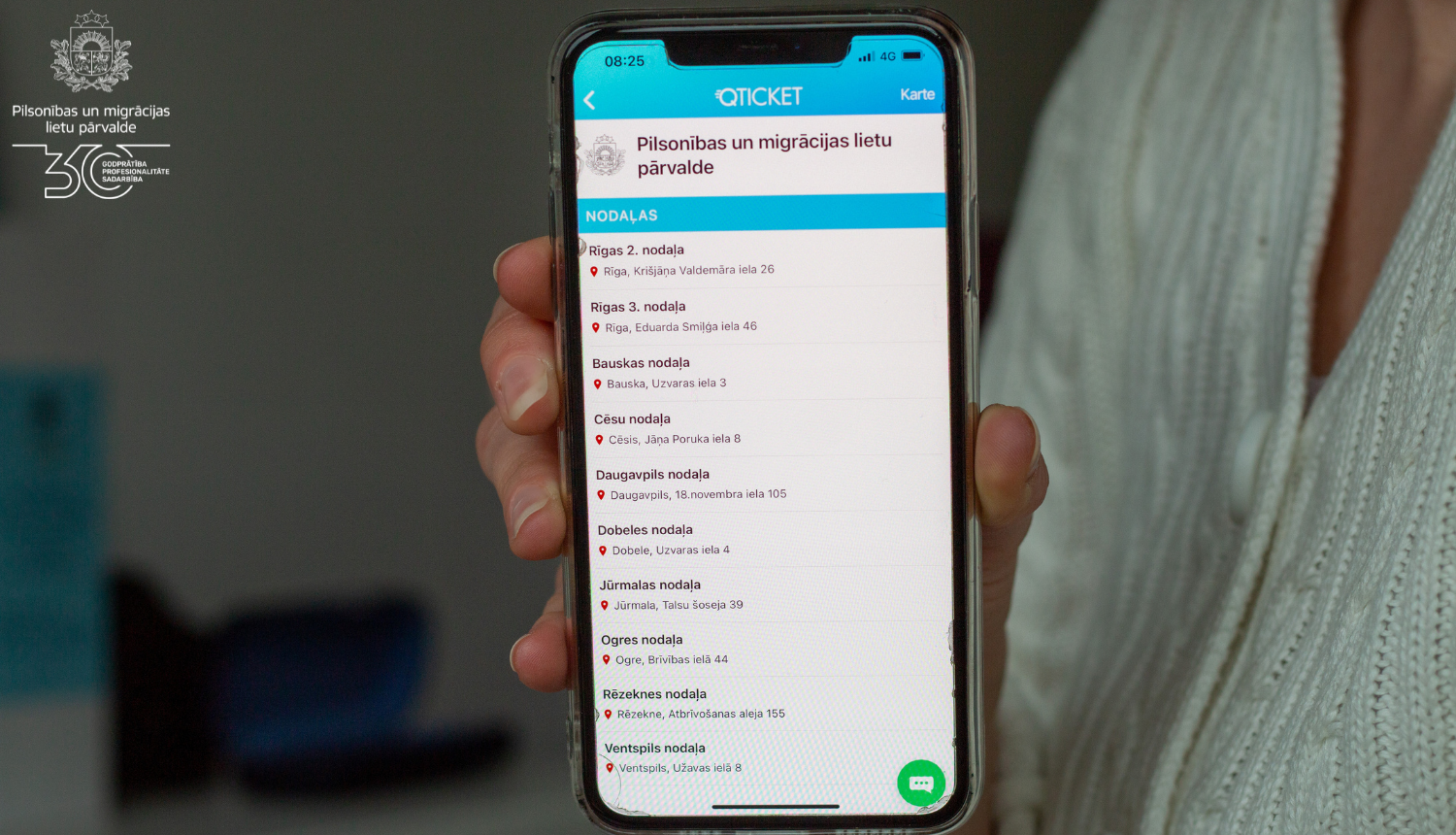 Sieviete tur rokā ar mobilo telefonu, kurā atvērta iepriekšējā pieraksta aplikācija Qticket, ar iespēju pierastīties vienā no PMLP nodaļām