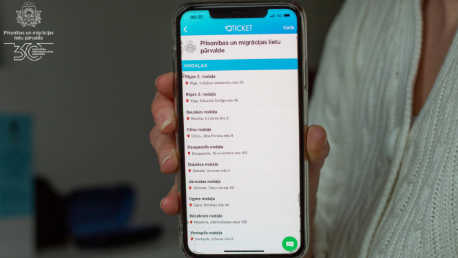 Sieviete tur rokā ar mobilo telefonu, kurā atvērta iepriekšējā pieraksta aplikācija Qticket, ar iespēju pierastīties vienā no PMLP nodaļām