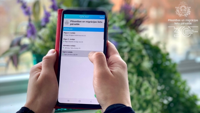 Cilvēka rokas, kas tur mobilo telefonu ar atvērtu Qticket aplikāciju un PMLP nodaļām, kurās iespējams veikt pierakstu