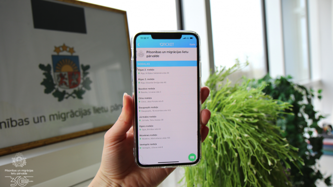 Cilvēks tur rokās telefonu ar Pārvaldes nodaļu sarakstu, kurās var veikt iepriekšējo pierakstu ar iepriekšējā pieraksta rīka starpniecību