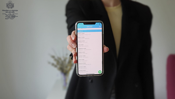 Sieviete melnā žaketē tur izstieptā rokā telefonu, ar atvērtu Qticket aplikāiju un iespēju pierakstīties vienā no PMLP nodaļām