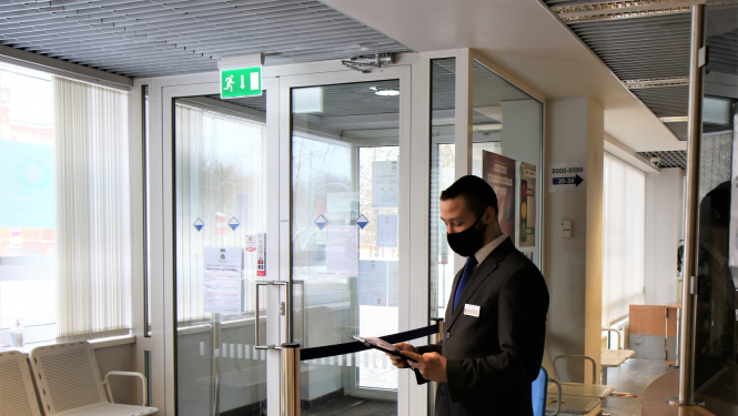 Klientu apkalpošanas speciālists ar uzvilktu mutes un deguna aizsegu un mapi rokās sagaida apmeklētājus pie Pārvades nodaļas ieejas durvīm