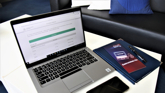 Uz galda: portatīvais dators ar atvērtu naturalizācijas pašpārbaudes e-rīku Pārvaldes mājaslapā,, mobilais telefons