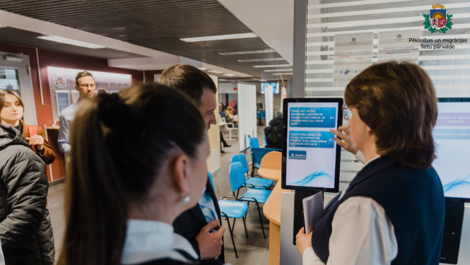 CIlvēki skatās uz pierakstu ekrānu, kur izsniedz numuriņus