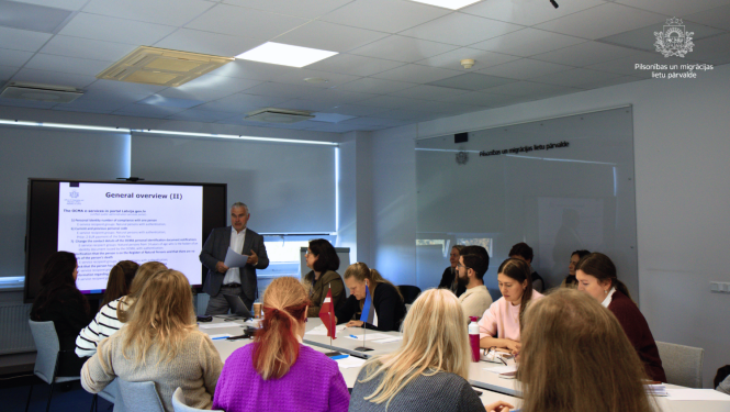 pie sanāksmes galda sēž vairāki sanāksmes dalībnieki un PMLP pārstāvis Uldis Apsītis uzstājas interaktīvas tāfeles priekšā