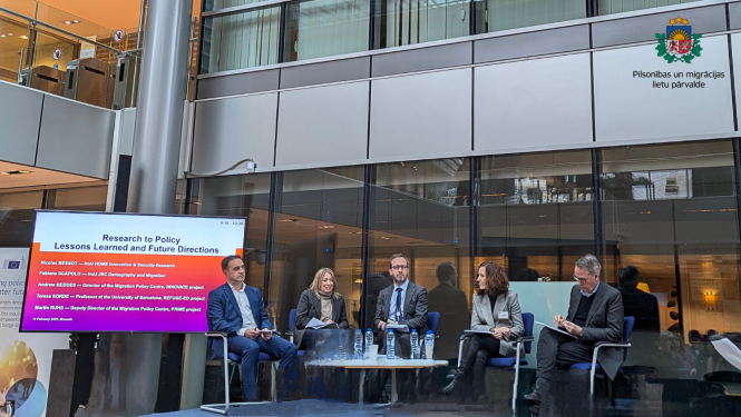 Cilvēki sēž uz skatuves un uzstājas, katrs tur rokās mikrofonus.