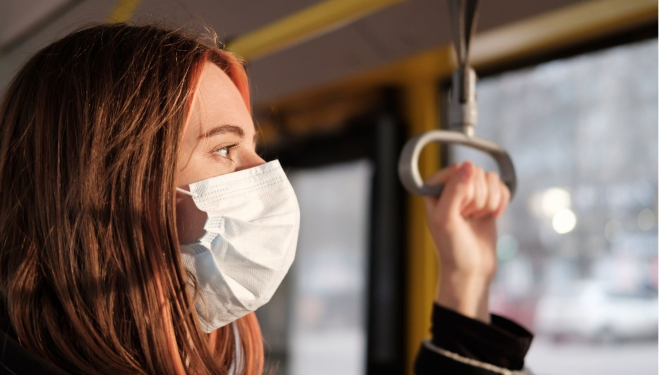 Jauna sieviete ar uzvilktu mutes un deguna aizsegu brauc sabiedriskajā transportā, skatās pa logu, 