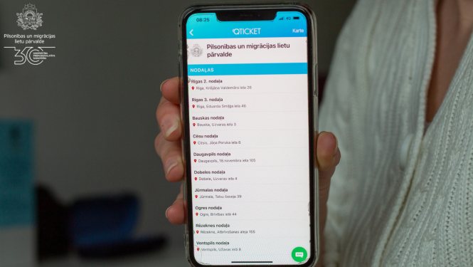 Sieviete tur rokā ar mobilo telefonu, kurā atvērta iepriekšējā pieraksta aplikācija Qticket, ar iespēju pierastīties vienā no PMLP nodaļām