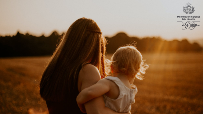 Mamma ar mazu bērnu rokās stāv ar muguru pret fotogrāfu saules pieļetajā pļāvā