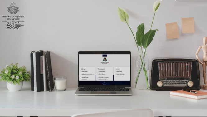 Uz galda stāv dators, pierakstu klades, vāzē - puķes