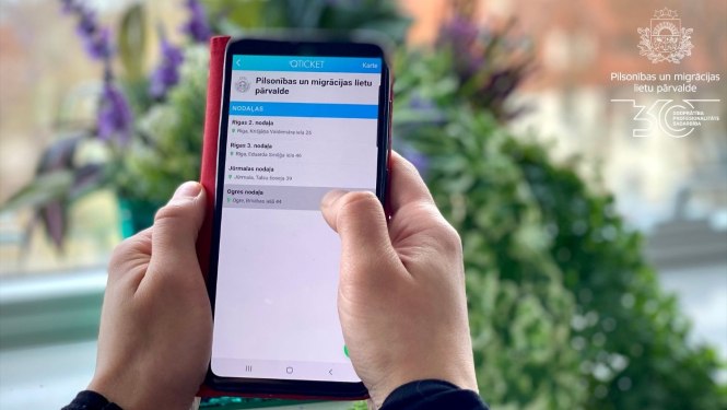 Cilvēka rokas, kas tur mobilo telefonu ar atvērtu Qticket aplikāciju un PMLP nodaļām, kurās iespējams veikt pierakstu