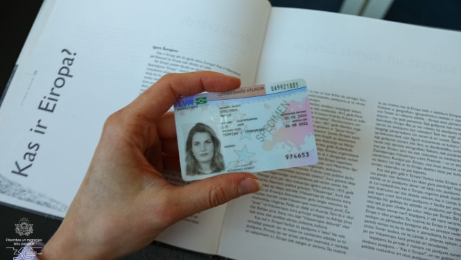 CIlvēka roka tur eID karti, uz grāmatas fona. Grāmatā nodaļas nosaukums "Kas ir Eiropa?"