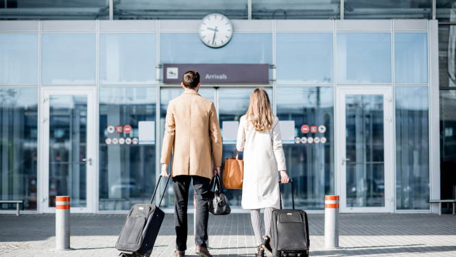 Vīrietis un sieviete stāv lidostas durvju priekšā, rokās ir rokās ir čemodāni