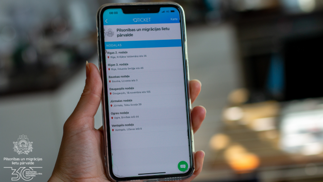 Cilvēks rokās tur mobilo telefonu ar atvērtu Qticket aplikāciju un iespēju pierakstīties vienā no 7 PMLP nodaļām
