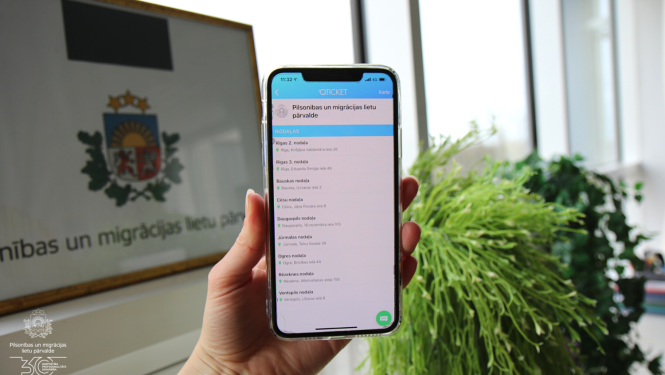 Cilvēks tur rokās telefonu ar Pārvaldes nodaļu sarakstu, kurās var veikt iepriekšējo pierakstu ar iepriekšējā pieraksta rīka starpniecību