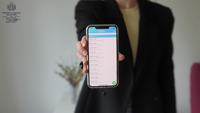 Sieviete melnā žaketē tur izstieptā rokā telefonu, ar atvērtu Qticket aplikāiju un iespēju pierakstīties vienā no PMLP nodaļām