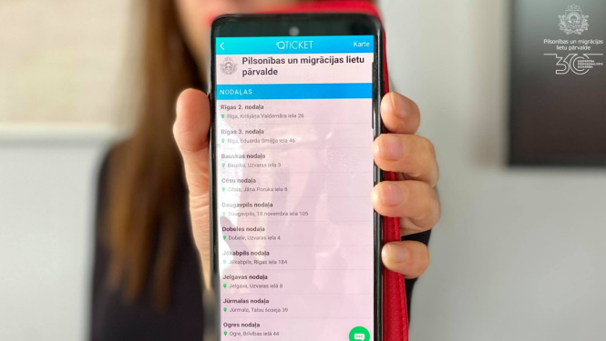 Sieviete melnā žaketē tur izstieptā rokā telefonu, ar atvērtu Qticket aplikāiju un iespēju pierakstīties vienā no PMLP nodaļām