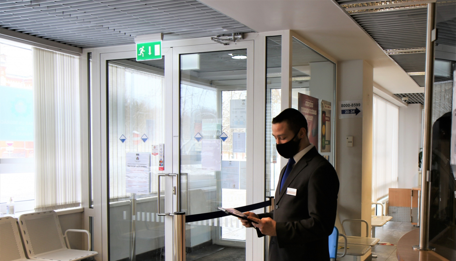 Klientu apkalpošanas speciālists ar uzvilktu mutes un deguna aizsegu un mapi rokās sagaida apmeklētājus pie Pārvades nodaļas ieejas durvīm