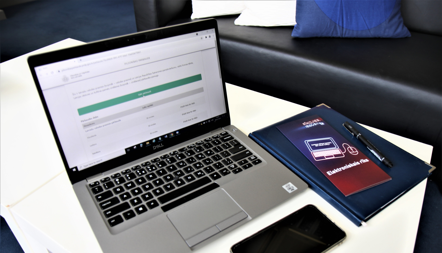Uz galda: portatīvais dators ar atvērtu naturalizācijas pašpārbaudes e-rīku Pārvaldes mājaslapā,, mobilais telefons