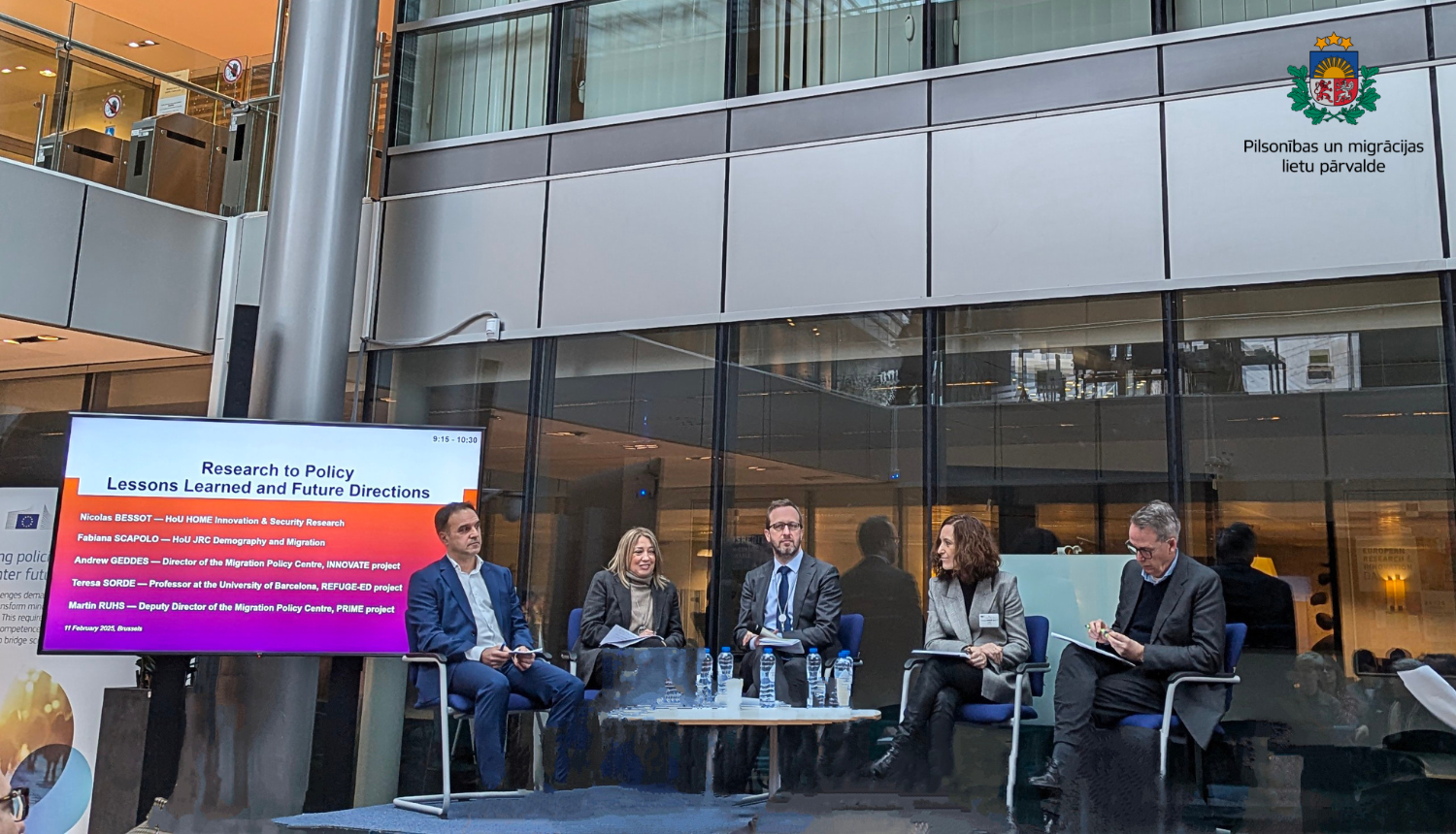 Cilvēki sēž uz skatuves un uzstājas, katrs tur rokās mikrofonus.