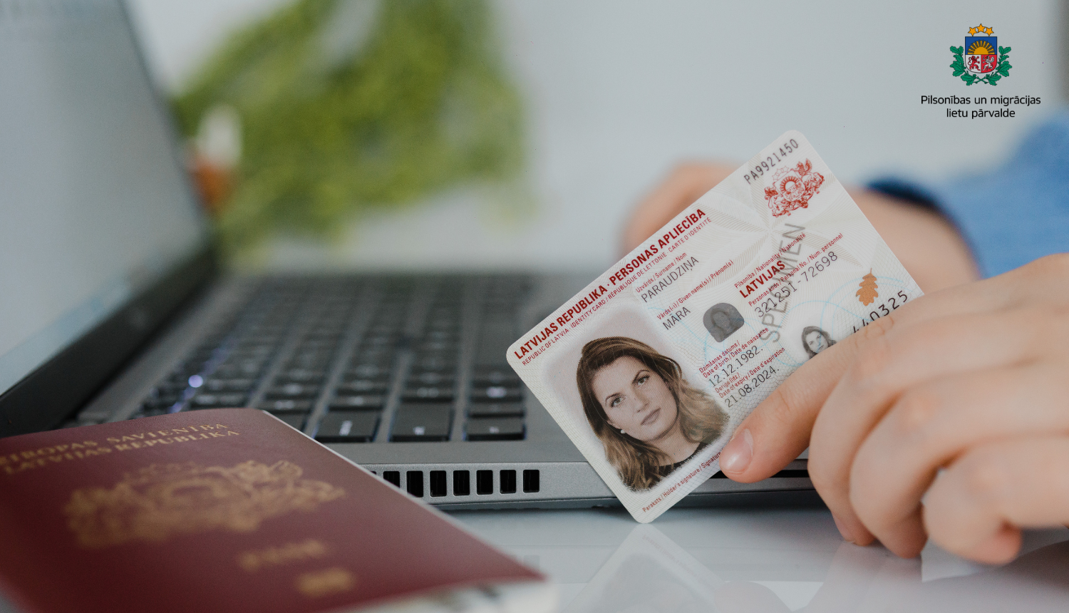 cilvēka roka  pie portatīvā datora tur eID karti u blakus uz galda - pase