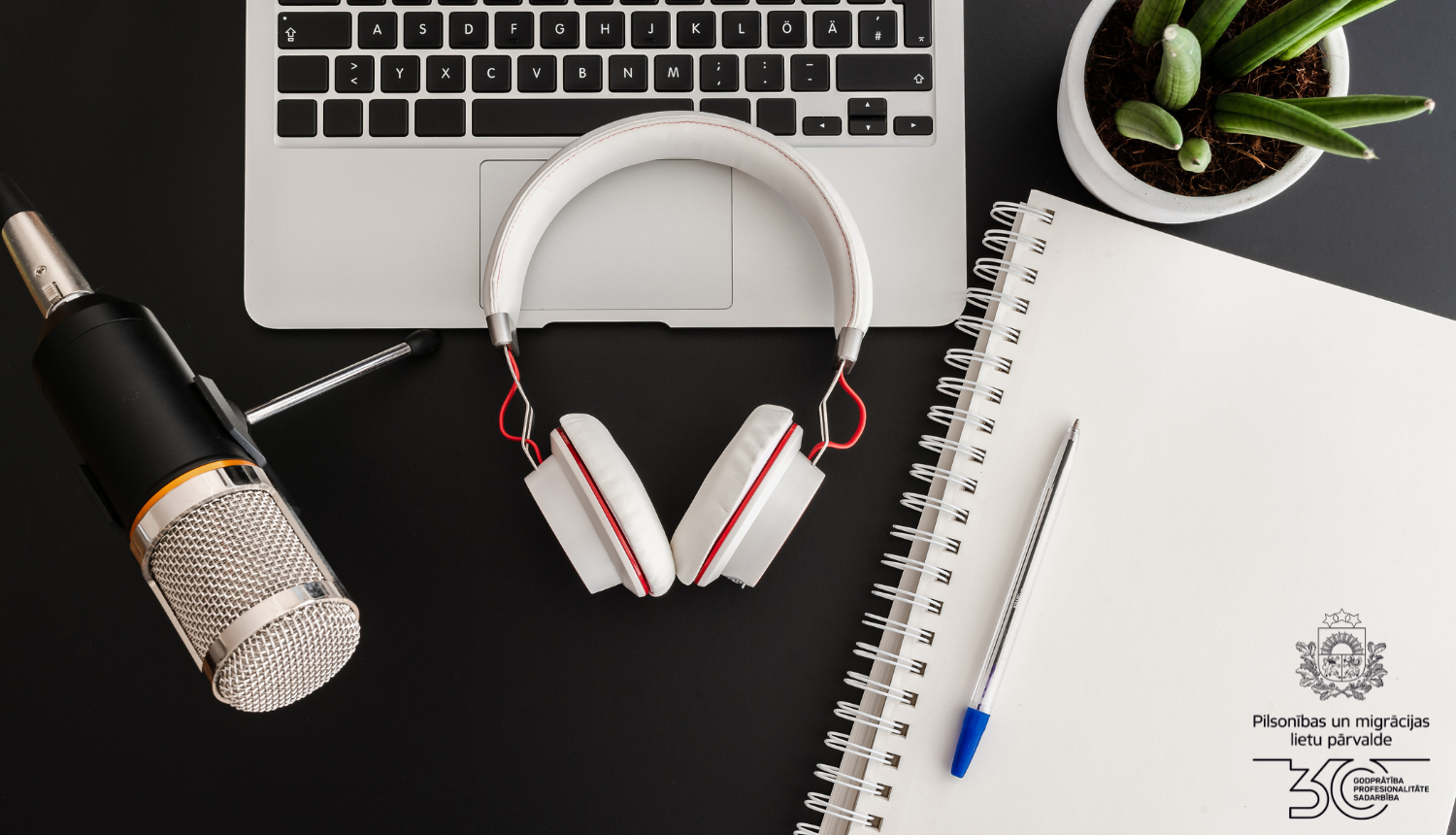 uz galda: austiņasmikrofons, portatīvais dators, pierakstu klade