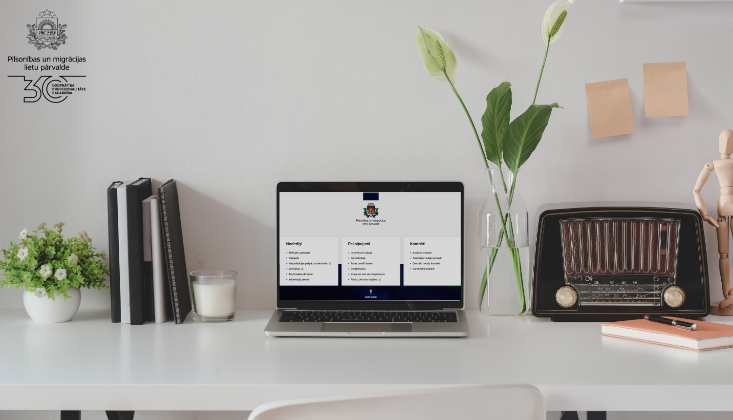 Uz galda stāv dators, pierakstu klades, vāzē - puķes