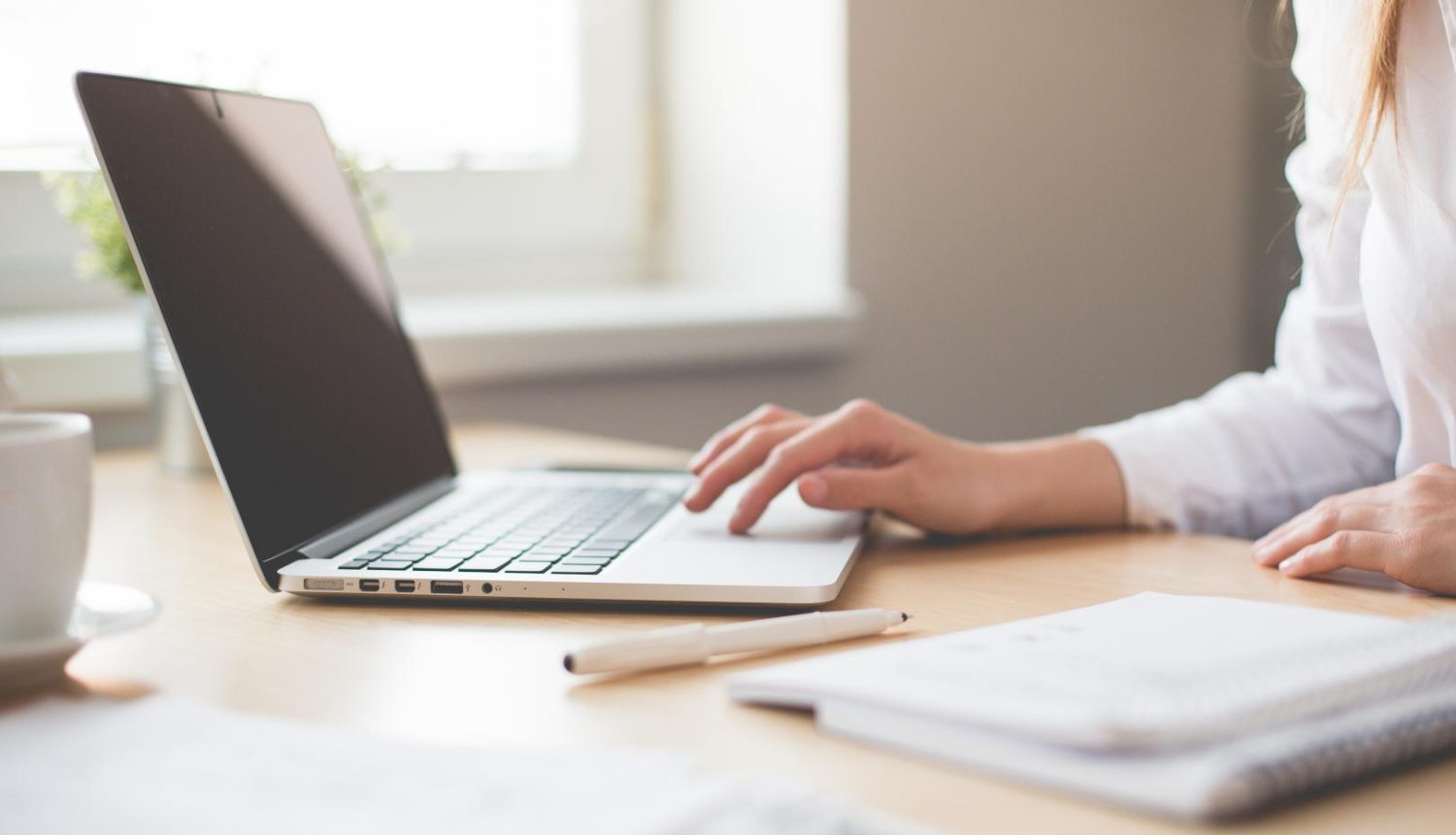 Sievietes rokas, lietojot portatīvo datoru, sēžot pie galda