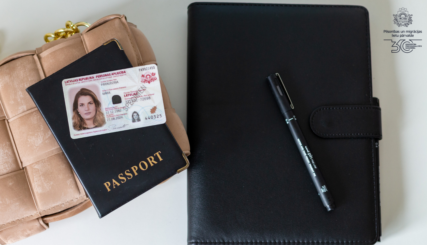 Uz galda: pierakstu klade melnajos vākos ar melnu pildspalvu, bēšīgās krāsas soma, un uz tās - pase melnajos vākos ar uzrakstu "Passport" un jaunā parauga ārzemnieku eID karte