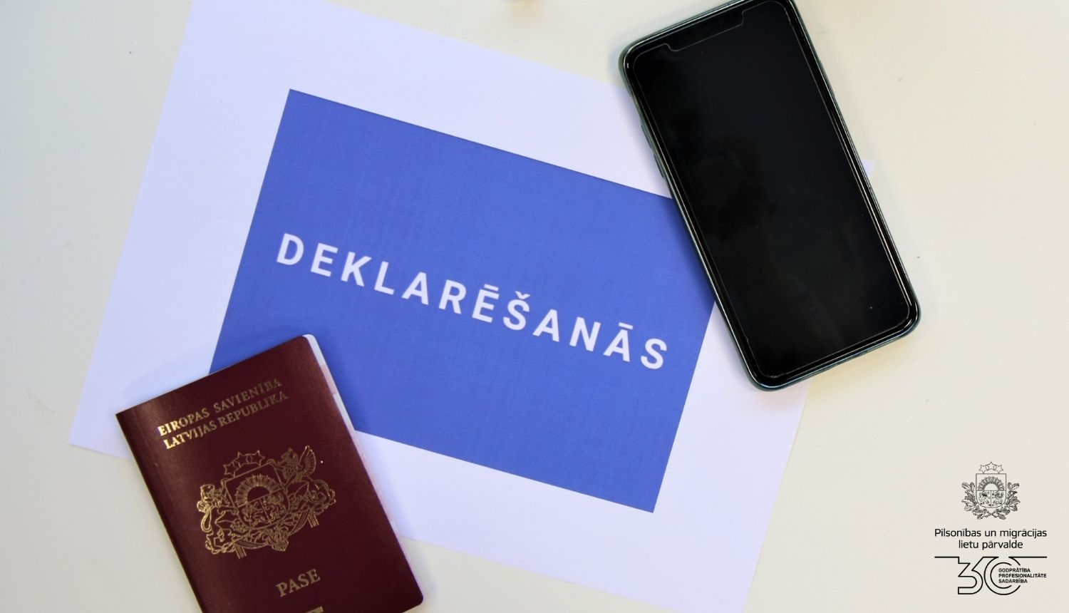 Kompozīcija uz balta galda: Latvijas pilsoņa pase, viedtālrunis un uzraksts uz zilā fona "Deklarēšanās"