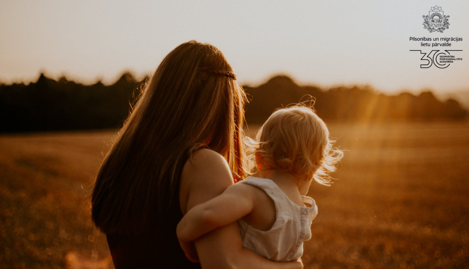 Mamma ar mazu bērnu rokās stāv ar muguru pret fotogrāfu saules pieļetajā pļāvā