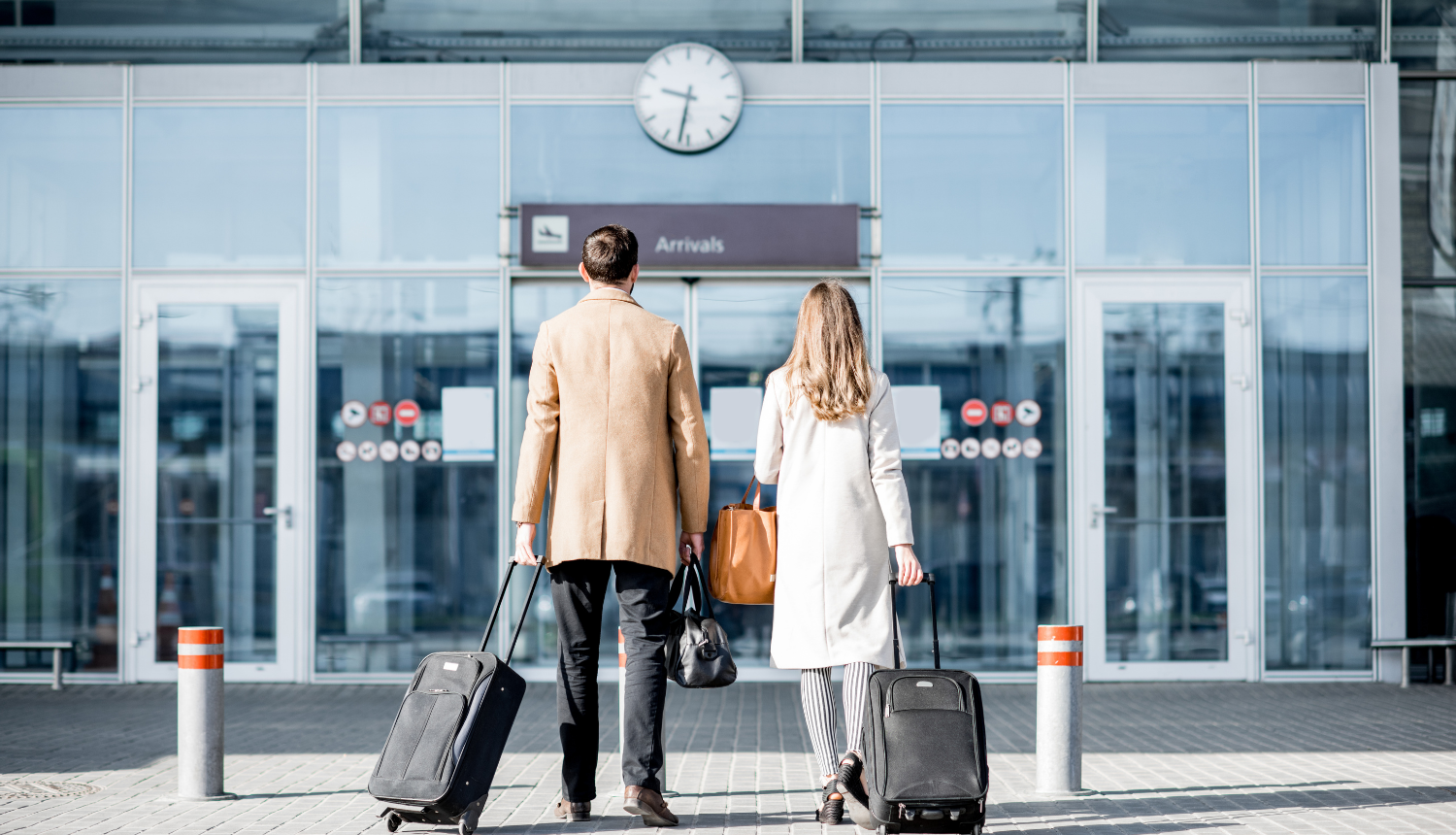 Vīrietis un sieviete stāv lidostas durvju priekšā, rokās ir rokās ir čemodāni
