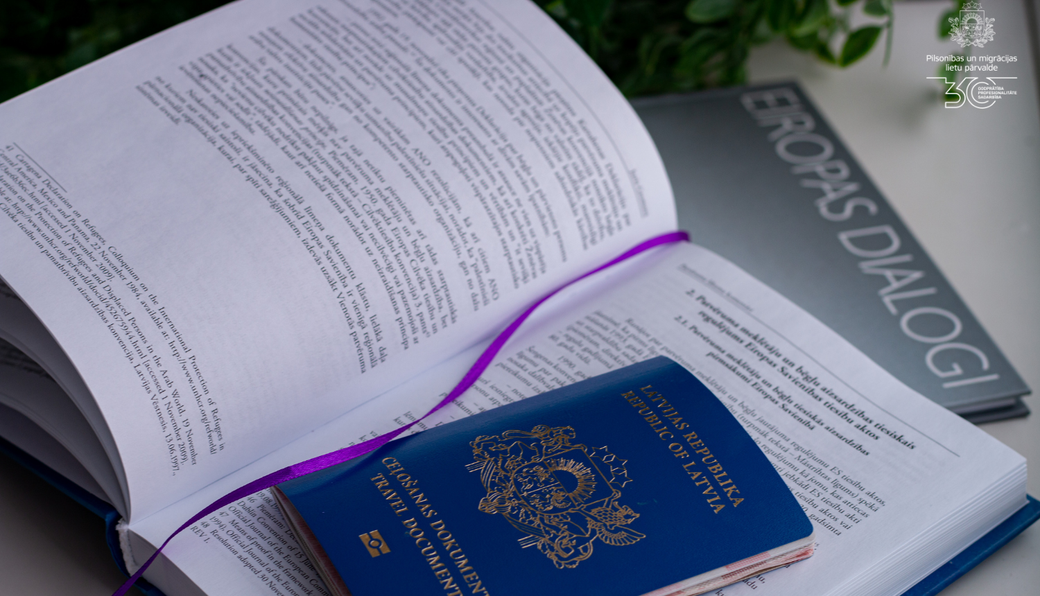 Atvērta grāmata, tajā ielikta zilā pase ar nosaukumu "Ceļošanas dokuments"