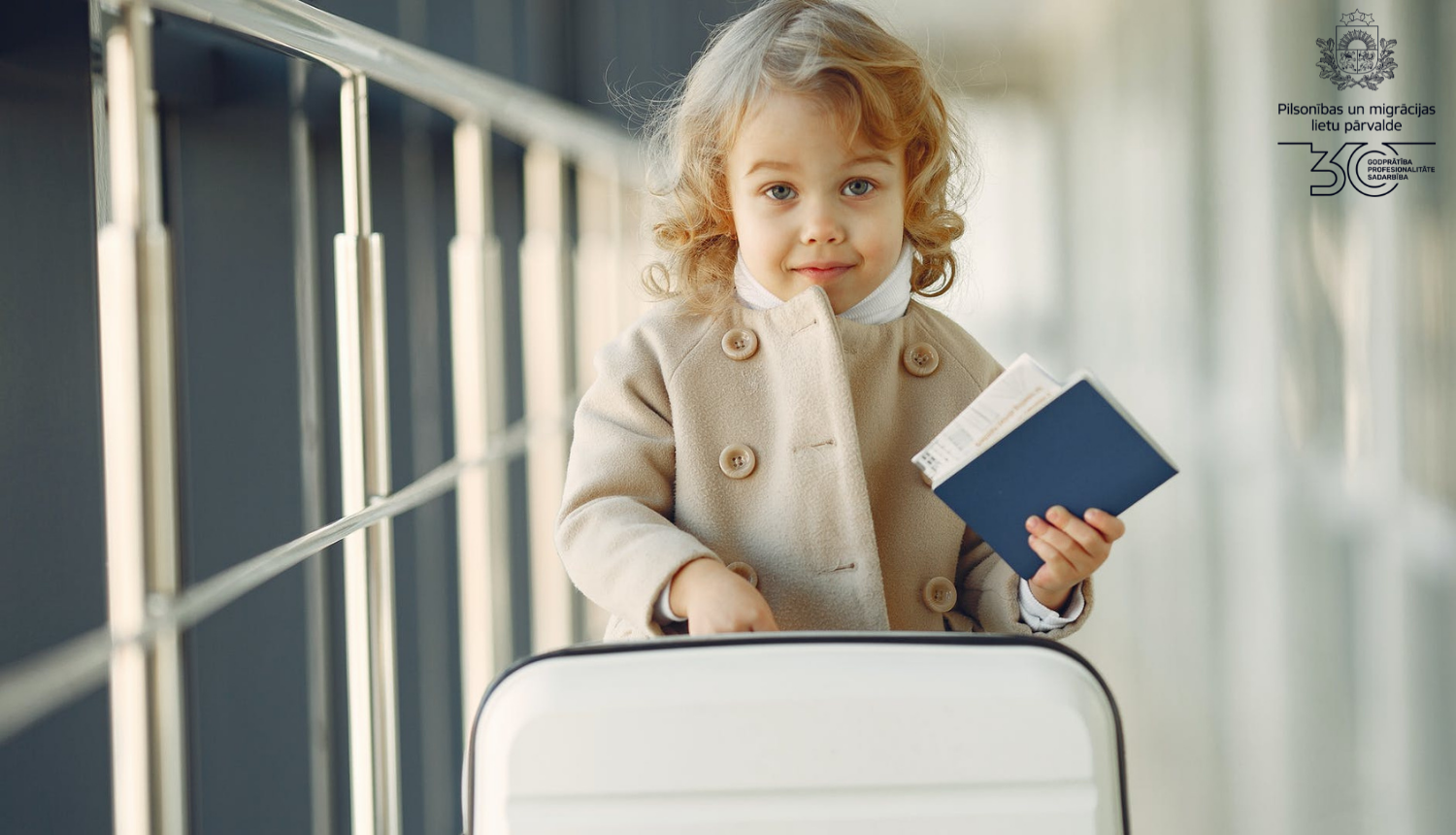 Mazs bērns stāv blakus koferim ar papīriem rokās