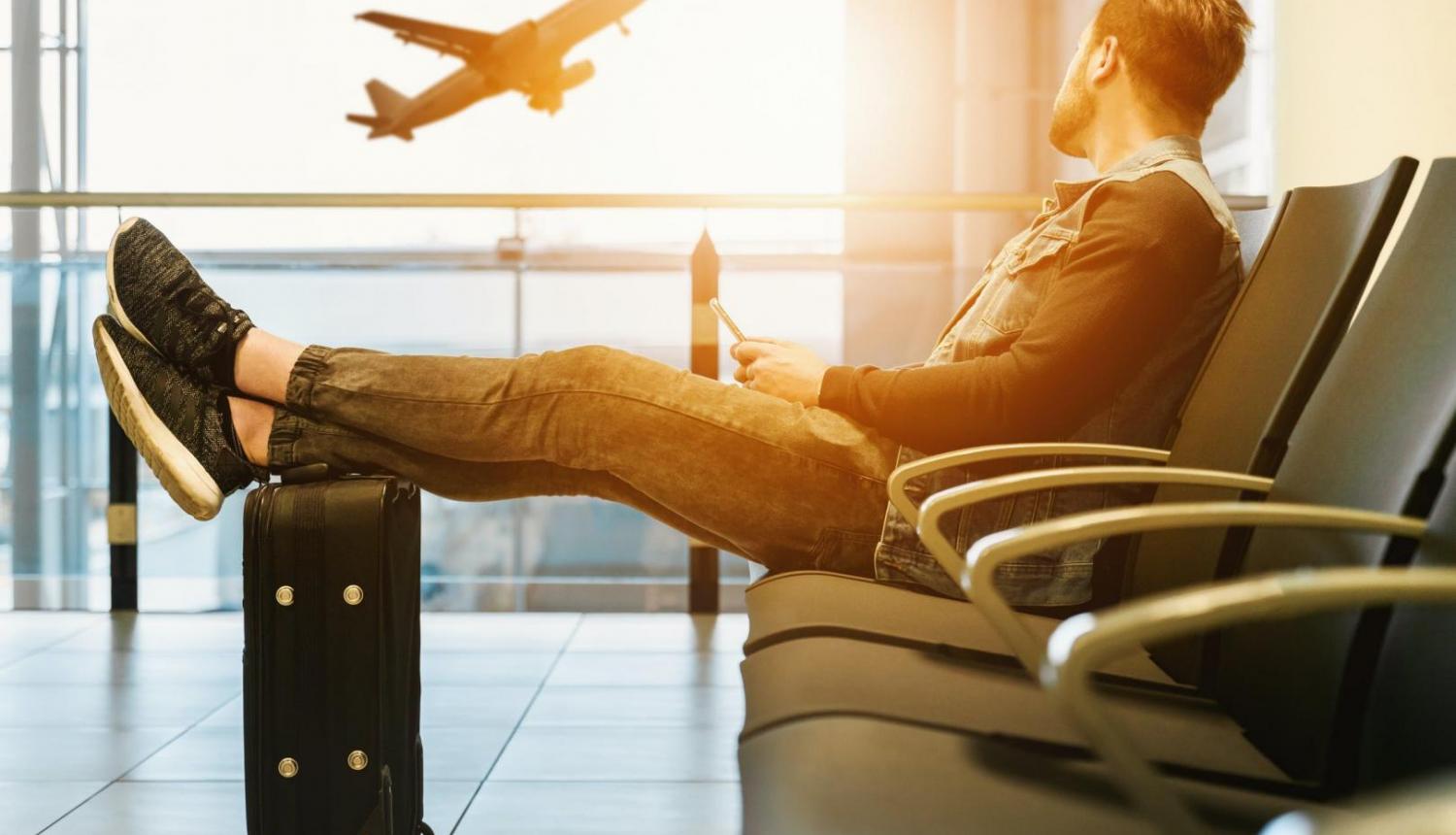 Vīrietis sēž lidostas uzgaidāmajā telpā uzlicis kādas uz čemodāna, skatās uz aizlidojošu lidmašīnu pa logu